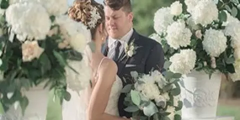 A bride and groom at a wedding surrounded by white floral arrangements and holding a bouquet.