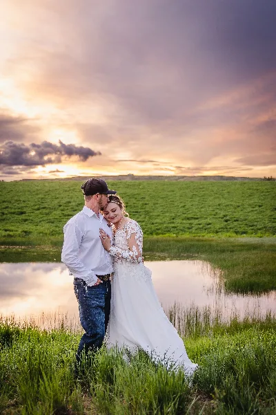 A bride and groom embracing in a grassy field near a body of water during a sunset.