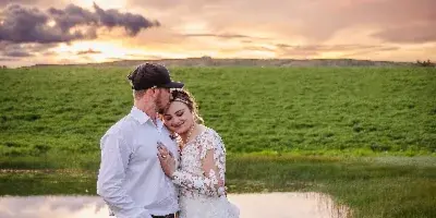 A bride and groom embracing in a grassy field near a body of water during a sunset.