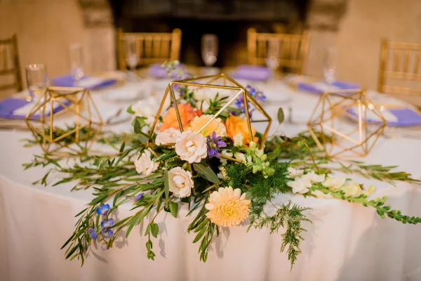 A wedding table centerpiece featuring a mix of white and peach flowers, greenery, and gold geometric frames on a white tablecloth.