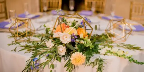 A wedding table centerpiece featuring a mix of white and peach flowers, greenery, and gold geometric frames on a white tablecloth.