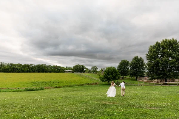 A bride and groom walking hand-in-hand across a green grassy field under a cloudy sky.