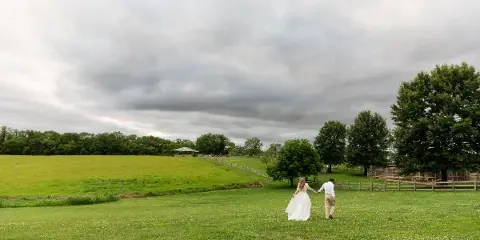 A bride and groom walking hand-in-hand across a green grassy field under a cloudy sky.
