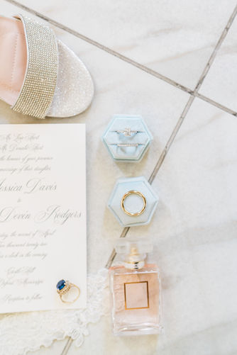 A flat lay photograph of wedding details including a ring box, invitation, perfume bottle, and a beaded shoe.