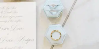 A flat lay photograph of wedding details including a ring box, invitation, perfume bottle, and a beaded shoe.