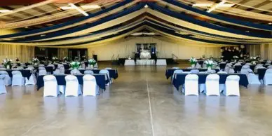 A spacious indoor event hall with round tables set for a wedding reception, featuring white chair covers and floral centerpieces under a high ceiling.