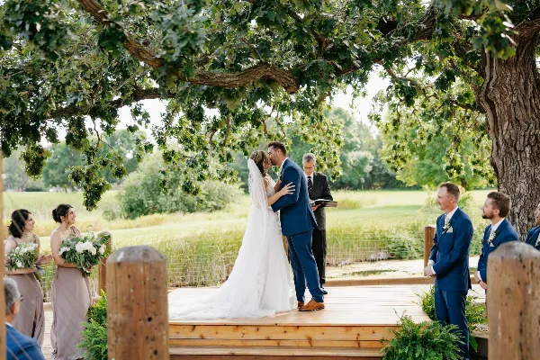 A bride and groom sharing a kiss during an outdoor wedding ceremony on a wooden stage beneath a large oak tree, surrounded by wedding party members.