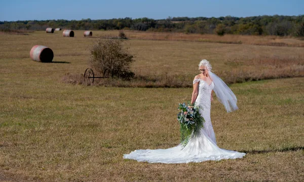 A bride in a white lace gown and veil holding a bouquet in a grassy field with hay bales in the background.