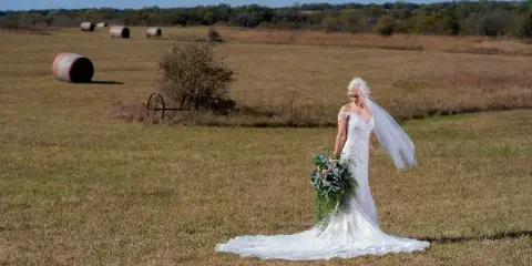 A bride in a white lace gown and veil holding a bouquet in a grassy field with hay bales in the background.