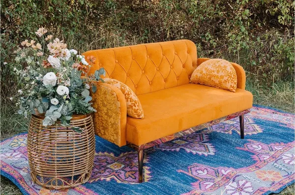 A mustard yellow velvet sofa with patterned pillows sits on a blue patterned rug next to a wicker stand holding a floral arrangement.