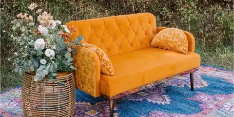 A mustard yellow velvet sofa with patterned pillows sits on a blue patterned rug next to a wicker stand holding a floral arrangement.