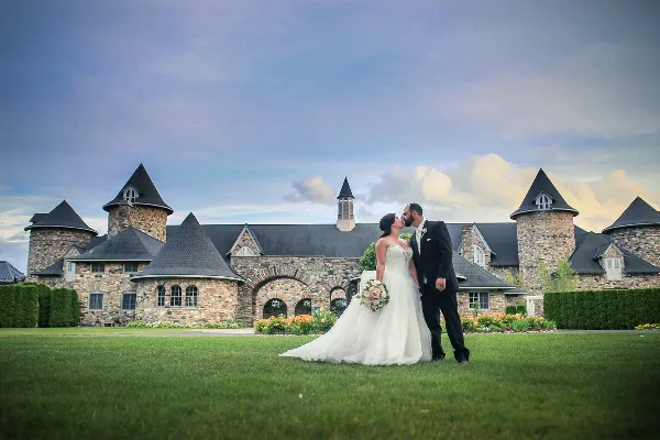A bride and groom kissing on a lawn in front of a large stone building with turrets.