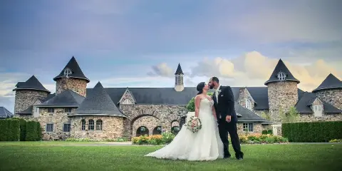 A bride and groom kissing on a lawn in front of a large stone building with turrets.