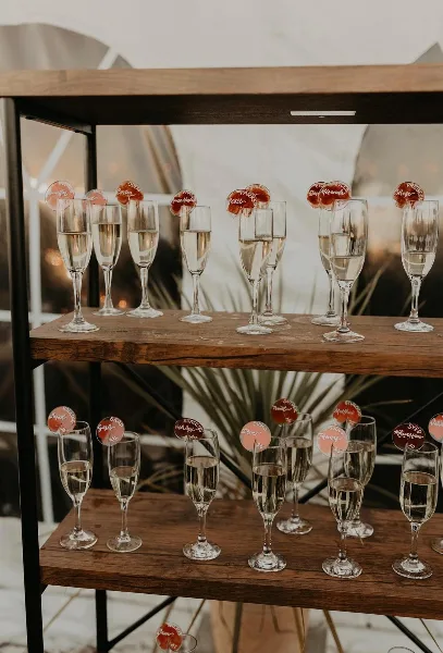 A wooden serving cart holding rows of champagne flutes with personalized name tags, set against a white backdrop.