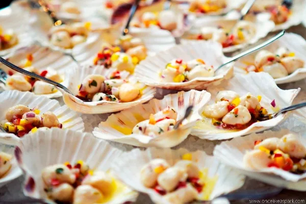 A close-up photograph of scallops topped with a colorful vegetable salsa, served in their shells.