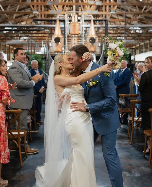 A bride and groom kiss during a wedding ceremony, with the bride holding a large, colorful bouquet of flowers.