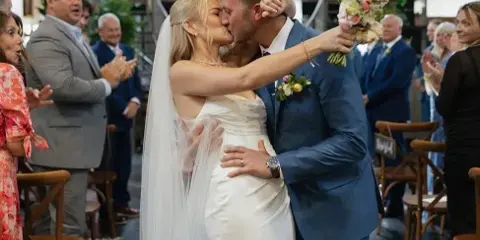 A bride and groom kiss during a wedding ceremony, with the bride holding a large, colorful bouquet of flowers.