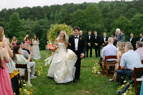 A bride and groom walking down an outdoor aisle with a floral arch and bouquet.
