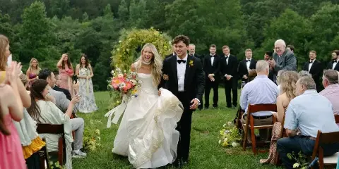 A bride and groom walking down an outdoor aisle with a floral arch and bouquet.