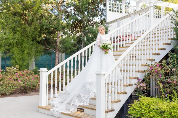 A bride in a white lace wedding dress and veil stands on a white staircase holding a bouquet of white flowers.