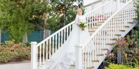 A bride in a white lace wedding dress and veil stands on a white staircase holding a bouquet of white flowers.
