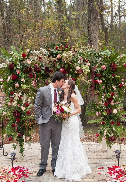 A bride and groom kissing in front of a lush floral wedding arch with red and pink roses and greenery.