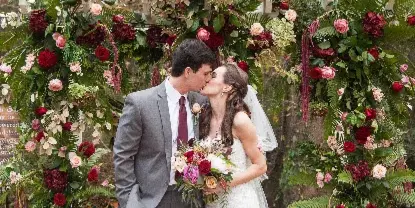 A bride and groom kissing in front of a lush floral wedding arch with red and pink roses and greenery.