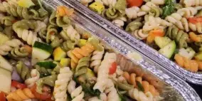 Large aluminum catering trays filled with tri-color rotini pasta salad mixed with vegetables.