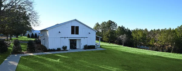 A white modern farmhouse event barn on a green lawn with a pond and trees in the background.