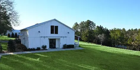 A white modern farmhouse event barn on a green lawn with a pond and trees in the background.