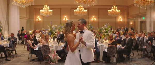 A bride and groom dancing in a large, elegant ballroom with chandeliers and seated guests.