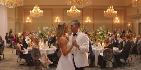 A bride and groom dancing in a large, elegant ballroom with chandeliers and seated guests.