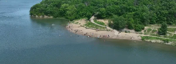 Aerial view of a park lake with a sandy shoreline, trees, and people recreating near the water.