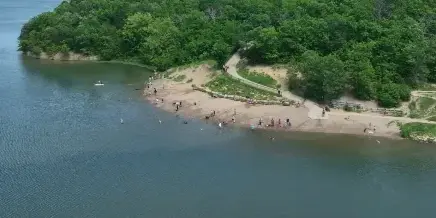 Aerial view of a park lake with a sandy shoreline, trees, and people recreating near the water.