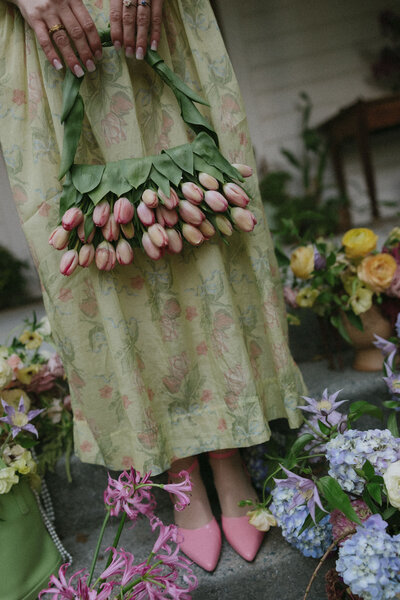 A close-up of a handcrafted floral basket made of pink tulips and green leaves, held by a person wearing a floral dress.