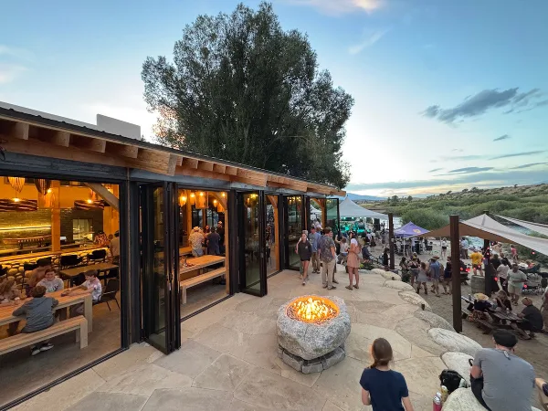 An indoor/outdoor venue with a stone patio, fire pit, and large glass doors opening to a dining area, set against a backdrop of a river and mountains.