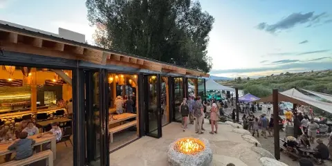 An indoor/outdoor venue with a stone patio, fire pit, and large glass doors opening to a dining area, set against a backdrop of a river and mountains.