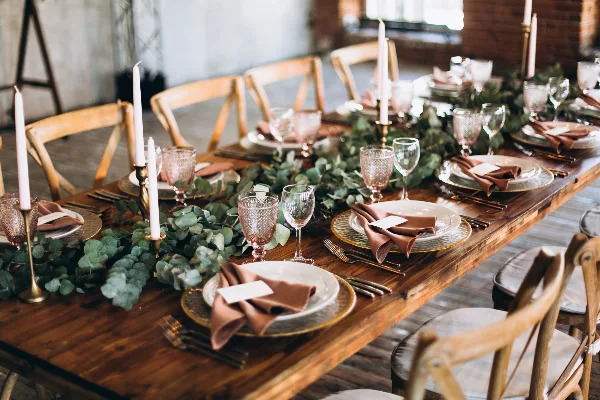 A long wooden table set for a wedding reception with a lush greenery centerpiece, tall white candles, and gold-rimmed plates.