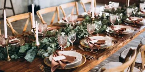 A long wooden table set for a wedding reception with a lush greenery centerpiece, tall white candles, and gold-rimmed plates.