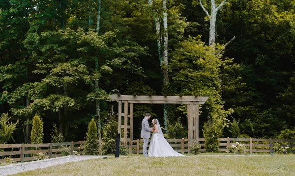 A bride and groom standing together beneath a wooden ceremony arbor in a grassy outdoor area surrounded by trees.