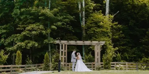 A bride and groom standing together beneath a wooden ceremony arbor in a grassy outdoor area surrounded by trees.