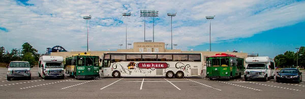 A lineup of Arkansas Destinations transportation vehicles, including a large white tour bus, green trolleys, and passenger vans, parked in a lot.
