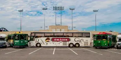 A lineup of Arkansas Destinations transportation vehicles, including a large white tour bus, green trolleys, and passenger vans, parked in a lot.