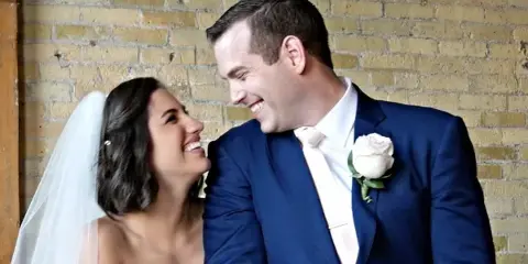 A bride and groom smiling at each other in front of a brick wall