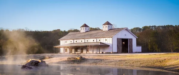 A large white barn with two cupolas and a covered porch situated on a farm property near a body of water.