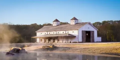 A large white barn with two cupolas and a covered porch situated on a farm property near a body of water.