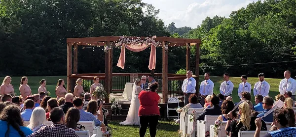 Outdoor wedding ceremony at a rustic venue with a wooden gazebo, floral decorations, and a green landscape backdrop.
