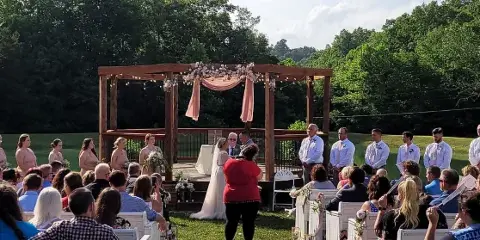 Outdoor wedding ceremony at a rustic venue with a wooden gazebo, floral decorations, and a green landscape backdrop.