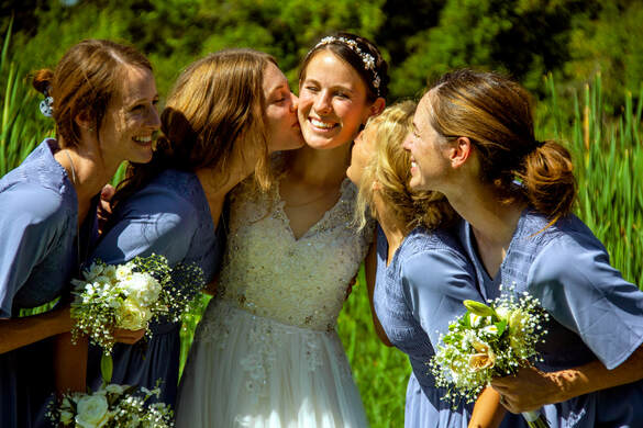 A bride in a white lace dress and a group of bridesmaids in blue dresses smiling and laughing together outdoors.
