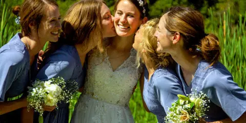 A bride in a white lace dress and a group of bridesmaids in blue dresses smiling and laughing together outdoors.